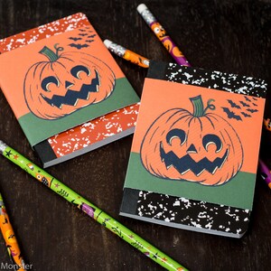 Può includere: Due piccoli quaderni a tema Halloween con copertine arancioni e verdi. Ogni quaderno presenta un'illustrazione in bianco e nero di una zucca con un ghigno spettrale. I quaderni sono circondati da matite colorate.