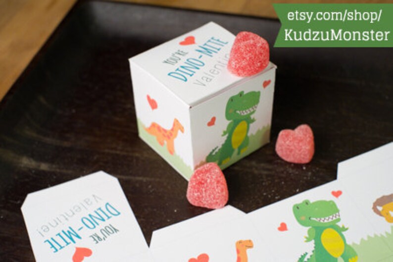 Puede incluir: Una caja blanca con un dise&ntilde;o de dinosaurio y el texto "You're Dino-Mite Valentine!" La caja est&aacute; llena de caramelos rojos en forma de coraz&oacute;n.