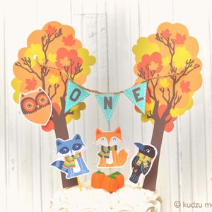 Puede incluir: Un adorno para pastel con dos árboles de otoño con hojas naranjas, amarillas y marrones. Los árboles están decorados con una pancarta que dice "ONE" en letras turquesa. También hay figuras de animales, incluyendo un búho, un zorro, un mapache y un cuervo, todos con bufandas. El adorno es perfecto para una fiesta de primer cumpleaños con temática de otoño.