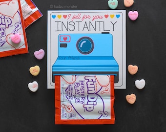 May include: A blue and white card with a camera design and the text "I fell for you INSTANTLY". The card has a red Fun Dip packet sticking out of the bottom. The card is surrounded by pink and white heart-shaped sweets.