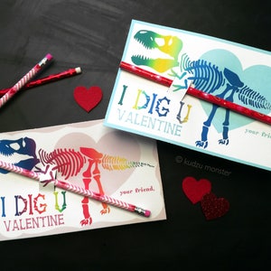 Könnte beinhalten: Zwei rosa und weiße Valentinstagskarten mit einem Regenbogen-Dinosaurierskelett-Motiv und dem Text "I DIG U VALENTINE". Jede Karte hat einen roten Bleistift mit dem Text "your friend" befestigt.