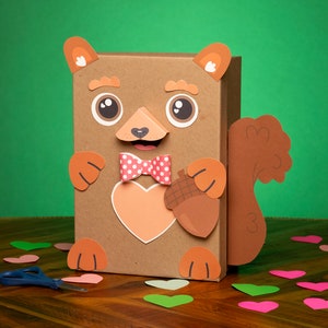 May include: A brown cardboard box decorated to look like a squirrel. The squirrel has a red bow tie with white polka dots, a brown acorn, and a heart-shaped cutout on its chest. The box is surrounded by pink and green paper hearts on a wooden surface.