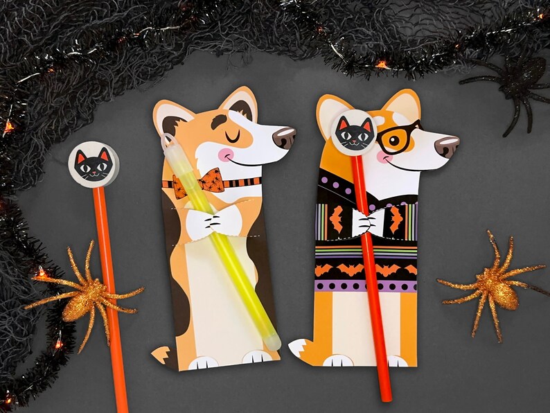 Op de afbeelding: Twee Halloween-potloodhouders in de vorm van corgis. E&eacute;n corgi draagt ​​een strikje en houdt een glowstick vast. De andere corgi draagt ​​een trui met vleermuizen en houdt een rood potlood vast.