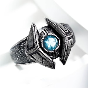 May include: A silver-toned ring with a central, light blue gemstone. The ring features a textured band and angular, decorative elements surrounding the gem. The design has a futuristic or fantasy aesthetic.