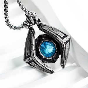May include: A silver-toned pendant necklace with a geometric design. The pendant features a central, round, blue gemstone. The necklace hangs on a silver chain, creating a striking contrast against the white background.