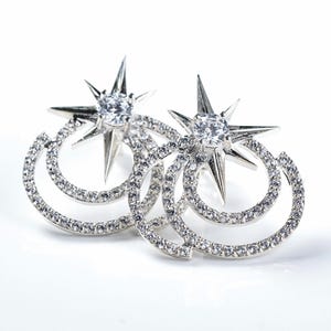 Könnte beinhalten: Ein Paar silberfarbene Ohrstecker. Jeder Ohrring hat ein sternförmiges Design mit einem zentralen, klaren Edelstein und einer doppelten Schlaufenform, die mit kleinen, klaren Kristallen besetzt ist. Auf weißem Hintergrund.