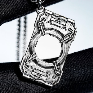 Puede incluir: Un colgante plateado con un diseño futurista e industrial. El colgante tiene forma rectangular con una gran abertura circular en el centro y detalles intrincados. Cuelga de una delicada cadena.