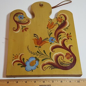 May include: A hand-painted wooden cutting board with a floral design in shades of blue, orange, yellow, and brown. The board has a hole at the top for hanging and is signed "Venice".