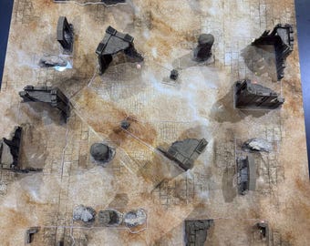 May include: A rectangular tabletop gaming mat featuring a tan and beige desert stone floor pattern with grid lines. The mat is populated with various grey, weathered stone ruin structures, including wall segments, pillars, and rocky debris.