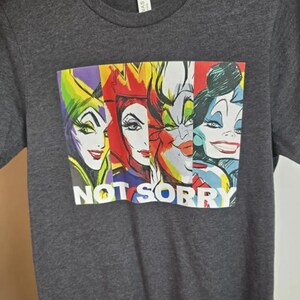 May include: Dark gray t-shirt featuring a colorful graphic of four cartoon villainesses. The graphic includes the text "NOT SORRY" in white block letters. The shirt is made of a soft material.