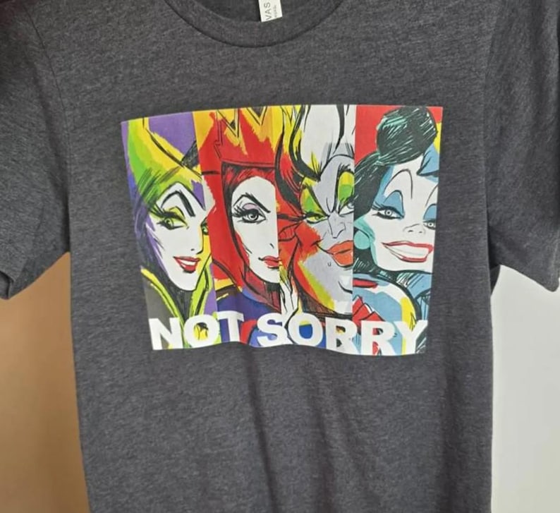 May include: Dark gray t-shirt featuring a colorful graphic of four cartoon villainesses. The graphic includes the text "NOT SORRY" in white block letters. The shirt is made of a soft material.
