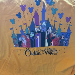 Puede incluir: Camiseta amarilla mostaza con un dise&ntilde;o de castillo, villanos de Disney y globos de Mickey Mouse. El texto "Chillin' Like a Villain" est&aacute; impreso debajo del castillo en una fuente estilizada.