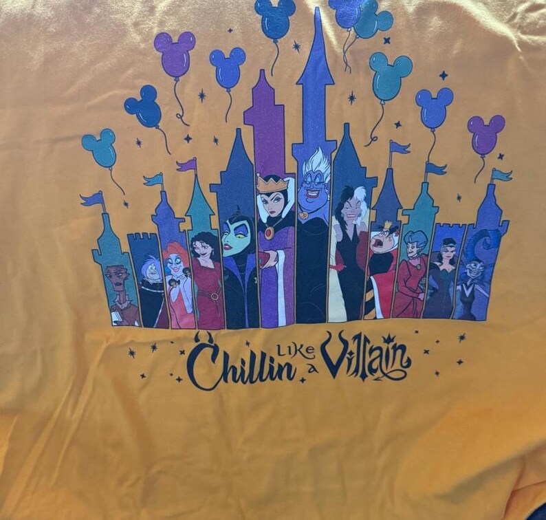 Puede incluir: Camiseta amarilla mostaza con un dise&ntilde;o de castillo, villanos de Disney y globos de Mickey Mouse. El texto "Chillin' Like a Villain" est&aacute; impreso debajo del castillo en una fuente estilizada.