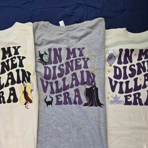 May include: Three t-shirts with the text "IN MY DISNEY VILLAIN ERA" in a retro font. Each shirt features a different Disney villain: Cruella de Vil, Maleficent, and Ursula. The shirts are cream and gray.