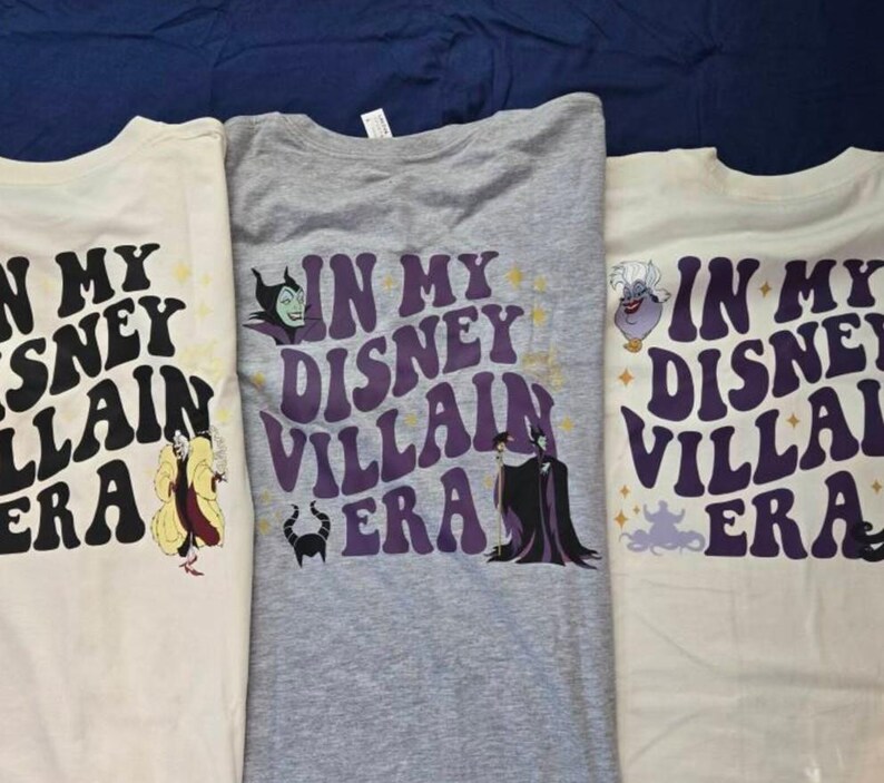 May include: Three t-shirts with the text "IN MY DISNEY VILLAIN ERA" in a retro font. Each shirt features a different Disney villain: Cruella de Vil, Maleficent, and Ursula. The shirts are cream and gray.