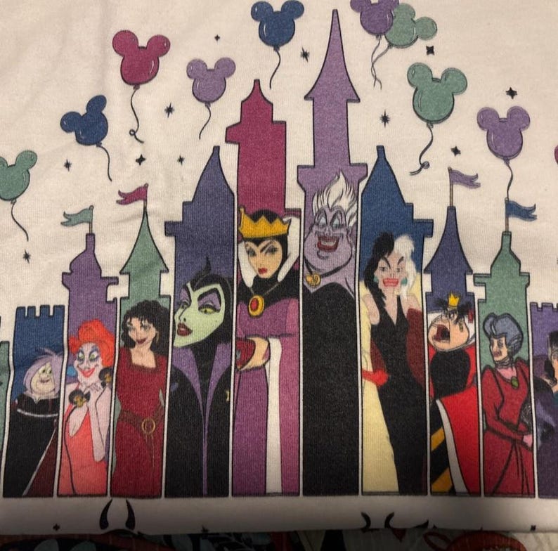 Puede incluir: Camiseta blanca con un gr&aacute;fico de villanos de Disney frente a una silueta de castillo. Globos con forma de Mickey Mouse flotan sobre el castillo. Los villanos incluyen a la Reina Malvada, &Uacute;rsula y Cruella de Vil.