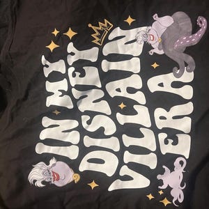 May include: Black t-shirt featuring the text "Disney Villain Era" in white, with a gold crown and star accents. The shirt includes illustrations of Ursula from The Little Mermaid.
