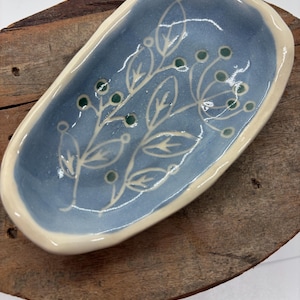 May include: A handmade ceramic soap dish. The dish is oval-shaped with a light blue interior and a cream-colored rim. It features a floral design in white and green. The dish is placed on a wooden surface.