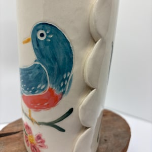 May include: A cylindrical ceramic vase with a glossy off-white finish, featuring a hand-painted blue bird with a red chest perched on a green branch with pink flowers. The side of the vase has a vertical row of raised, scalloped petal-like shapes.