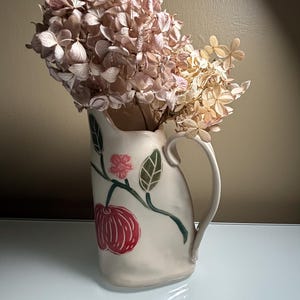 May include: A cream-colored ceramic pitcher with a curved handle, featuring a hand-painted design of a red apple, a small pink flower, and green leaves on a vine. The pitcher is filled with a large bouquet of dried, light brown hydrangea flowers.