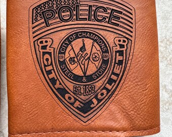 May include: A brown leatherette can cooler featuring a black engraved police badge design. The badge includes the word "POLICE" above an American flag, and the text "CITY OF CHAMPIONS JOLIET" with "STEEL & STONE" and "Est. 1852".