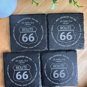 May include: Four square black slate coasters with the Route 66 logo, celebrating the 100th anniversary from 1926-2026. Each coaster features the iconic shield design with "ROUTE 66" and the mileage, 2,448 miles.