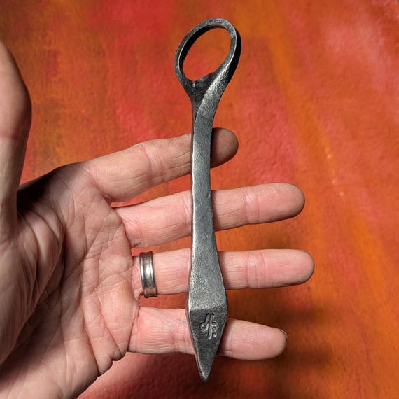 A blacksmith literally hammered this bottle opener from raw iron — the forge marks and hand-finished texture make it look like it belongs in a medieval tavern.