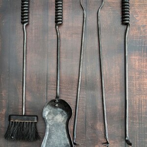 May include: A set of five black wrought iron fireplace tools hanging on a decorative metal bar. The tools include a brush, shovel, poker, tongs, and a curved tool, all with unique handle designs. The tools are displayed against a dark wood background.