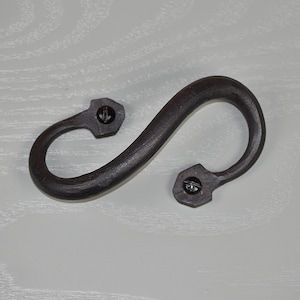 Op de afbeelding: Een donkerbruine, handgesmede ijzeren S-haak. De haak heeft een gebogen vorm met twee bevestigingspunten, elk met een schroef. Deze decoratieve hardware is geschikt voor rustieke of boerderij-inrichting.
