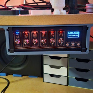 May include: A black and gray ELEKS USB SWITCH device with multiple toggle switches and a small display, mounted under a wooden shelf. The device is surrounded by other electronic components and small storage drawers.