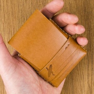 May include: A brown leather wallet with a stitched design and a small bird graphic. The wallet is folded in half and held in a hand.