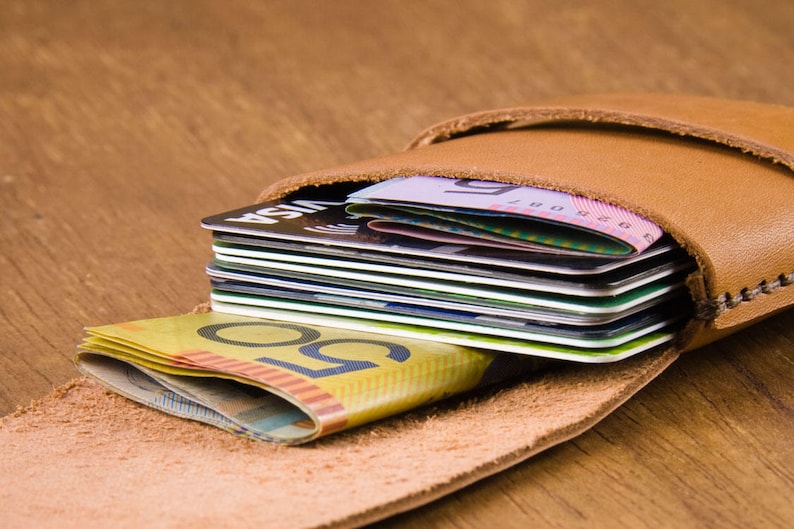 Puede incluir: Una cartera de cuero marr&oacute;n con cierre de solapa, abierta para revelar una pila de tarjetas de cr&eacute;dito y algunos billetes. La cartera est&aacute; sobre una superficie de madera.
