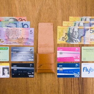 May include: A brown leather wallet with a variety of cards and Australian currency bills. The cards include a Medicare card, a platinum VISA debit card, a driver's license, a Sunshine Beach Country Club golf card, and a Flybugs VISA debit card. The bills include five, ten, twenty, and fifty Australian dollar bills.