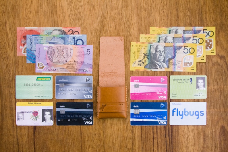 May include: A brown leather wallet with a variety of cards and Australian currency bills. The cards include a Medicare card, a platinum VISA debit card, a driver's license, a Sunshine Beach Country Club golf card, and a Flybugs VISA debit card. The bills include five, ten, twenty, and fifty Australian dollar bills.