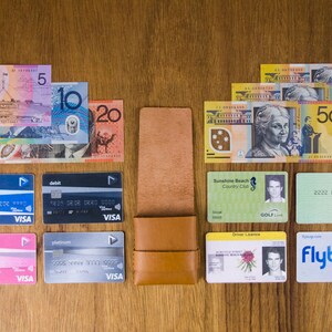 Puede incluir: Una cartera de cuero marr&oacute;n que contiene una variedad de tarjetas y billetes de banco australianos. Las tarjetas incluyen una tarjeta de d&eacute;bito, una tarjeta de cr&eacute;dito, un permiso de conducir, una tarjeta de membres&iacute;a de un club de golf, una tarjeta Medicare y una tarjeta Flybuys. Los billetes de banco son en denominaciones de 5, 10, 20 y 50 d&oacute;lares australianos.