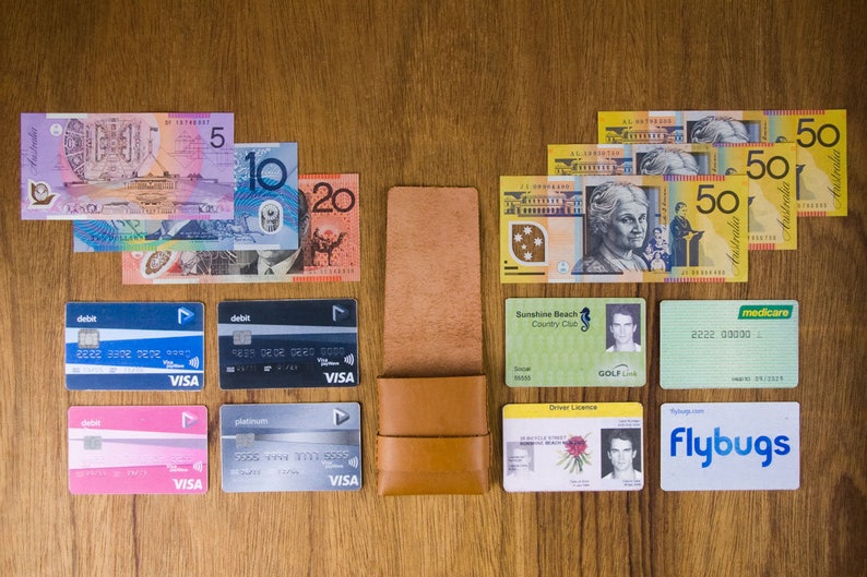 Puede incluir: Una cartera de cuero marr&oacute;n que contiene una variedad de tarjetas y billetes de banco australianos. Las tarjetas incluyen una tarjeta de d&eacute;bito, una tarjeta de cr&eacute;dito, un permiso de conducir, una tarjeta de membres&iacute;a de un club de golf, una tarjeta Medicare y una tarjeta Flybuys. Los billetes de banco son en denominaciones de 5, 10, 20 y 50 d&oacute;lares australianos.