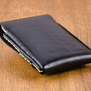 May include: Black leather wallet with a stitched edge and a cream colored stitching detail. The wallet is closed and lying on a wooden surface.