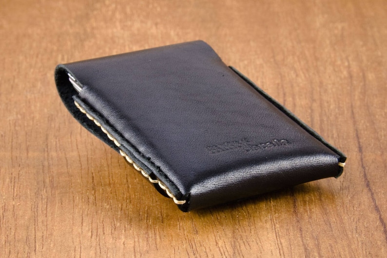 May include: Black leather wallet with a stitched edge and a cream colored stitching detail. The wallet is closed and lying on a wooden surface.