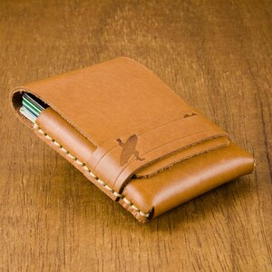 May include: A brown leather card holder with a stitched edge and a subtle design of a bird in flight.