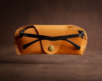May include: A tan leather glasses case with a snap closure and a pair of black glasses inside. The glasses have a black frame and black lenses.