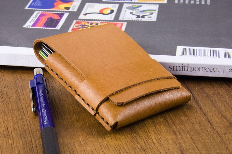 Puede incluir: Una cartera de cuero marr&oacute;n con un borde cosido y un cierre de solapa. La cartera est&aacute; sobre una superficie de madera.