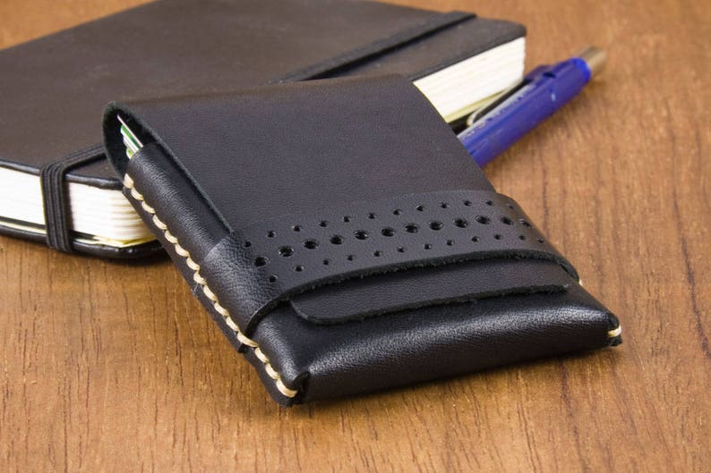 May include: A black leather wallet with a stitched design and a cream colored border. The wallet has a flap closure and is lying on a wooden surface.