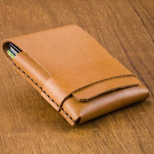 Puede incluir: Una cartera de cuero marr&oacute;n con un borde cosido. La cartera est&aacute; cerrada y se encuentra sobre una superficie de madera.