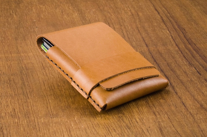 Puede incluir: Una cartera de cuero marr&oacute;n con un borde cosido. La cartera est&aacute; cerrada y se encuentra sobre una superficie de madera.