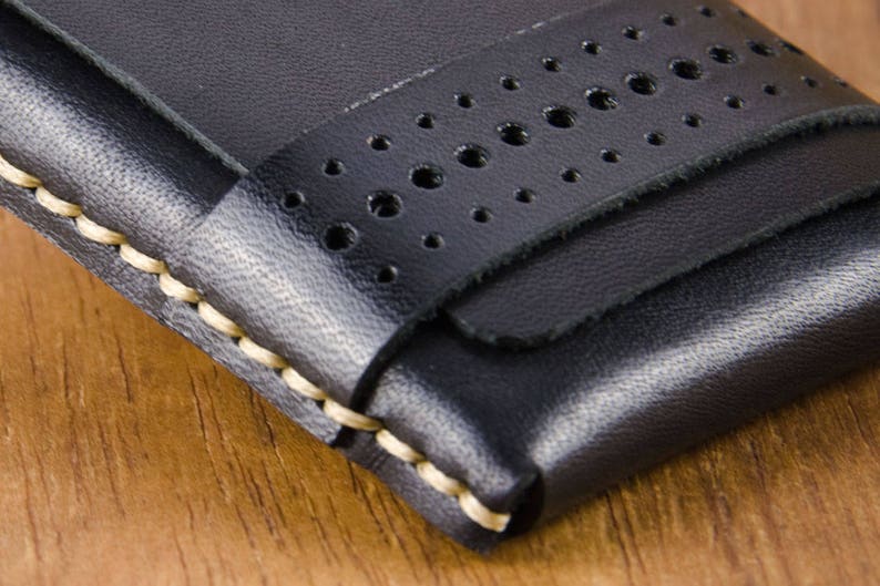 May include: A black leather wallet with a stitched edge and a perforated strap. The wallet has a minimalist design.