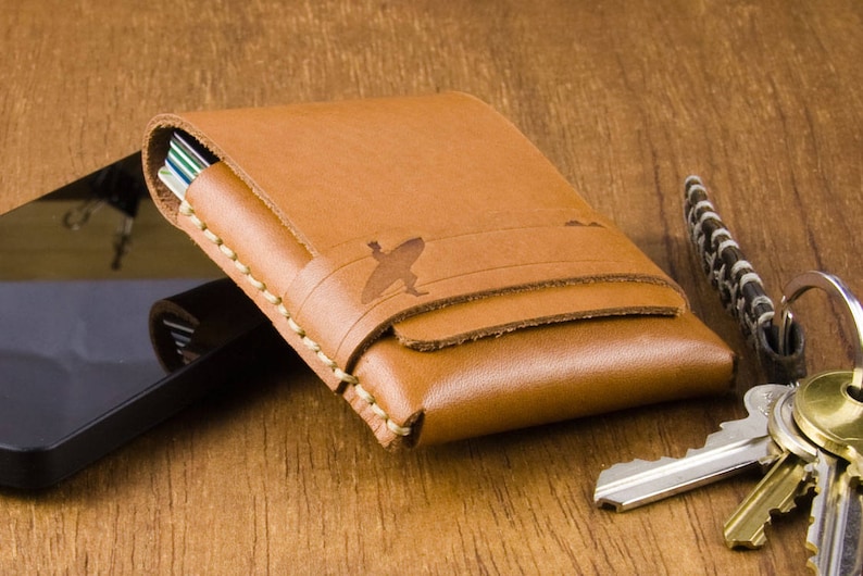 May include: A brown leather wallet with a stitched edge and a small design on the front. The wallet is closed and lying on a wooden surface.