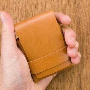 Puede incluir: Una cartera de cuero marr&oacute;n con una solapa plegable y una costura. La cartera se sostiene en una mano.
