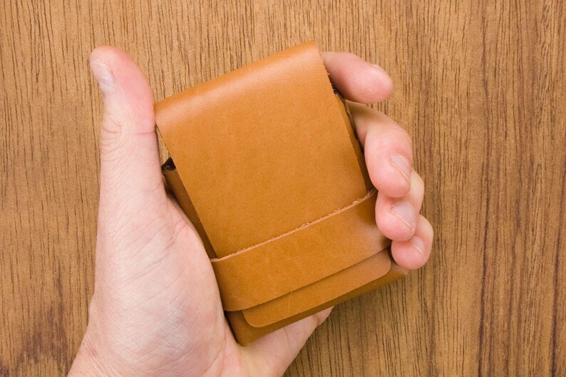 Puede incluir: Una cartera de cuero marr&oacute;n con una solapa plegable y una costura. La cartera se sostiene en una mano.