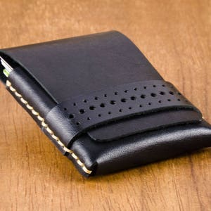 May include: A black leather card holder with a stitched edge and a perforated strap. The holder has a flap that closes with a snap.