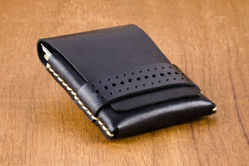 May include: A black leather card holder with a stitched edge and a perforated strap. The holder has a flap that closes with a snap.